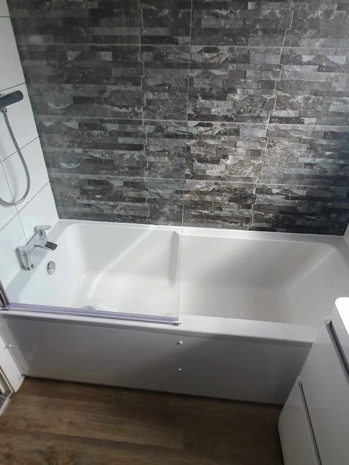 White bath with a glass shower screen and dark stone-effect wall tiles