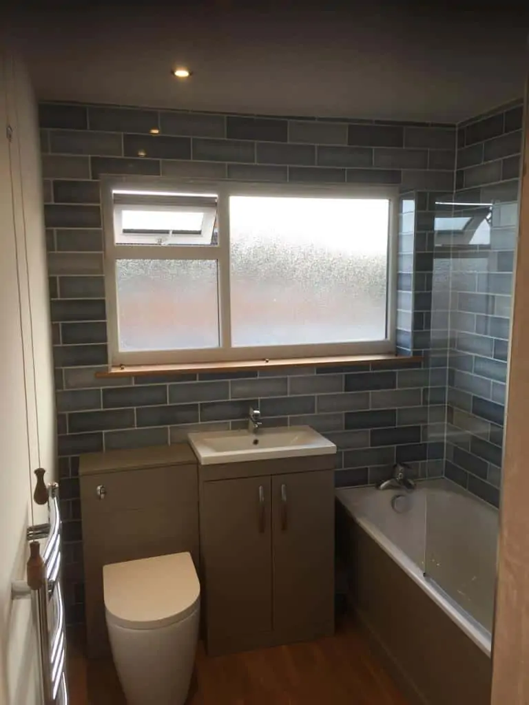 Bathroom with grey tiled walls, a vanity unit, toilet and bath beneath a frosted window