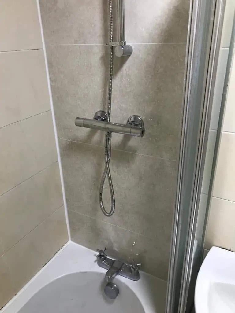Shower with a chrome mixer, handheld shower and bath taps against tiled walls.